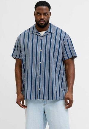 Mężczyzna o krótkich włosach i brodzie, ubrany w niebiesko-białą koszulę z pionowymi paskami na krótki rękaw zapinaną na guziki oraz jasnoniebieskie dżinsy.