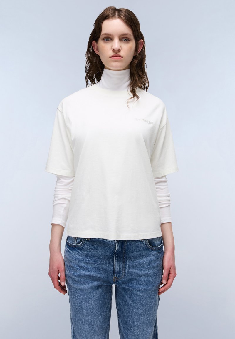 White cotton T-shirt with a high neck, short sleeves, and a small logo on the chest. Worn over a long-sleeve top and paired with blue jeans.