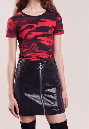 Femme portant une chemise à manches courtes imprimé camouflage rouge et noir rentrée dans une mini-jupe noire brillante zippée à l'avant avec une texture imitation peau de serpent.