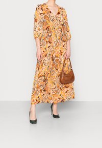 Długa, wzorzysta sukienka w odcieniach pomarańczowym, brązowym i zielonym, z dekoltem w kształcie litery V i rękawami trzyczwartowymi. Połączona z brązową torbą i zielonymi szpilkami.