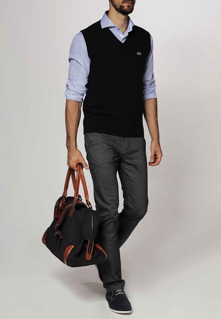 Svart ärmlös tröja över en ljusblå rutad skjorta, grå byxor och marinblå skor. Håller en svart väska med bruna detaljer.