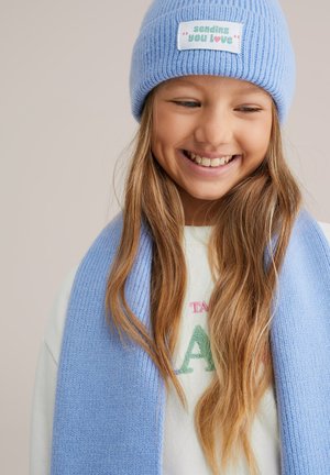 Jauna mergaitė šypsosi, dėvinti šviesiai mėlynę megztą kepurę su etikete „siunčiu tau meilę“ ir derančią skaros šaliką, uždėtą ant balto megztinio.