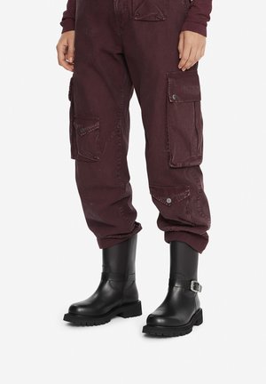 Persona che indossa pantaloni cargo color viola scuro con multiple tasche e stivali neri a metà polpaccio con fibbia laterale, in piedi su uno sfondo semplice.