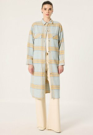 Une femme porte un long manteau à carreaux bleu pâle et jaune sur un pantalon crème, devant un fond clair uni.