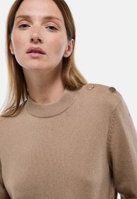 Femme aux cheveux châtain clair portant un pull en laine beige avec deux boutons décoratifs sur l'épaule, sur un fond clair uni.