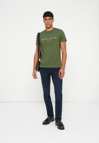 Grøn bomulds t-shirt med logo, rund halsudskæring og korte ærmer, parret med mørkeblå jeans og sorte loafers, bærende en taske.