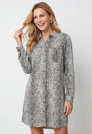 Lächelnde Frau mit langen blonden Haaren, die goldene Creolen und ein schwarz-weiß gepunktetes Hemdkleid trägt, berührt mit einer Hand ihr Haar.
