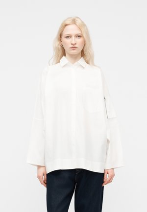 Young person with long blonde hair wearing a loose white button-up shirt with wide sleeves and dark pants, standing against a plain background.