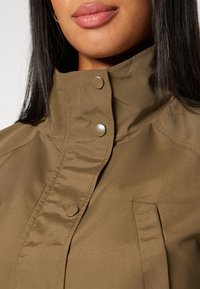 Giacca khaki con colletto alto, bottoni a pressione e una texture liscia. Presenta un dettaglio di tasca nascosta nella parte anteriore.