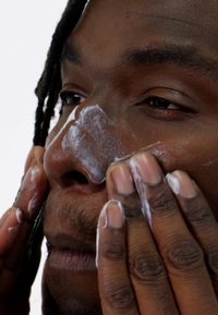 Routine de nettoyage du visage avec un nettoyant crémeux blanc appliqué sur un teint foncé, les doigts massant doucement le produit sur la peau.
