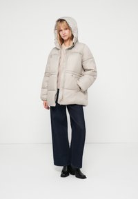 Chaqueta acolchada beige con capucha, que cuenta con un cierre de cremallera frontal, dos bolsillos delanteros y puños elásticos. Lleva una combinación con jeans oscuros de pierna ancha y botas negras.