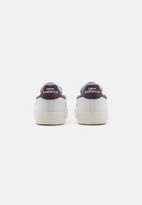 Fehér vászon sportcipő lila díszítéssel a sarkán, texturált felülettel és bézs gumitalppal. A saroknál a "New Balance" logó.