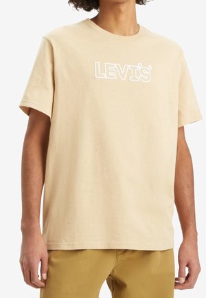 Beige Levi's t-shirt med rund hals, båret af en person med mellemtonet hud og krøllet hår, kombineret med sennepsfarvede bukser.