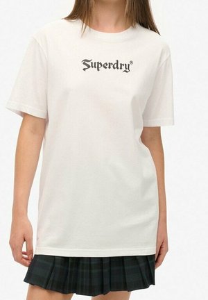 Kobieta ubrana w gładką białą koszulkę z krótkim rękawem z napisem „Superdry” i ciemnozieloną plisowaną spódnicę na białym tle.