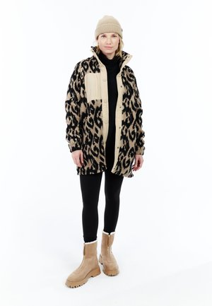 Vrouw in een beige beanie, een fleece jack met luipaardprint en tan accenten, zwarte legging en beige bontline laarzen, staand op een witte achtergrond.