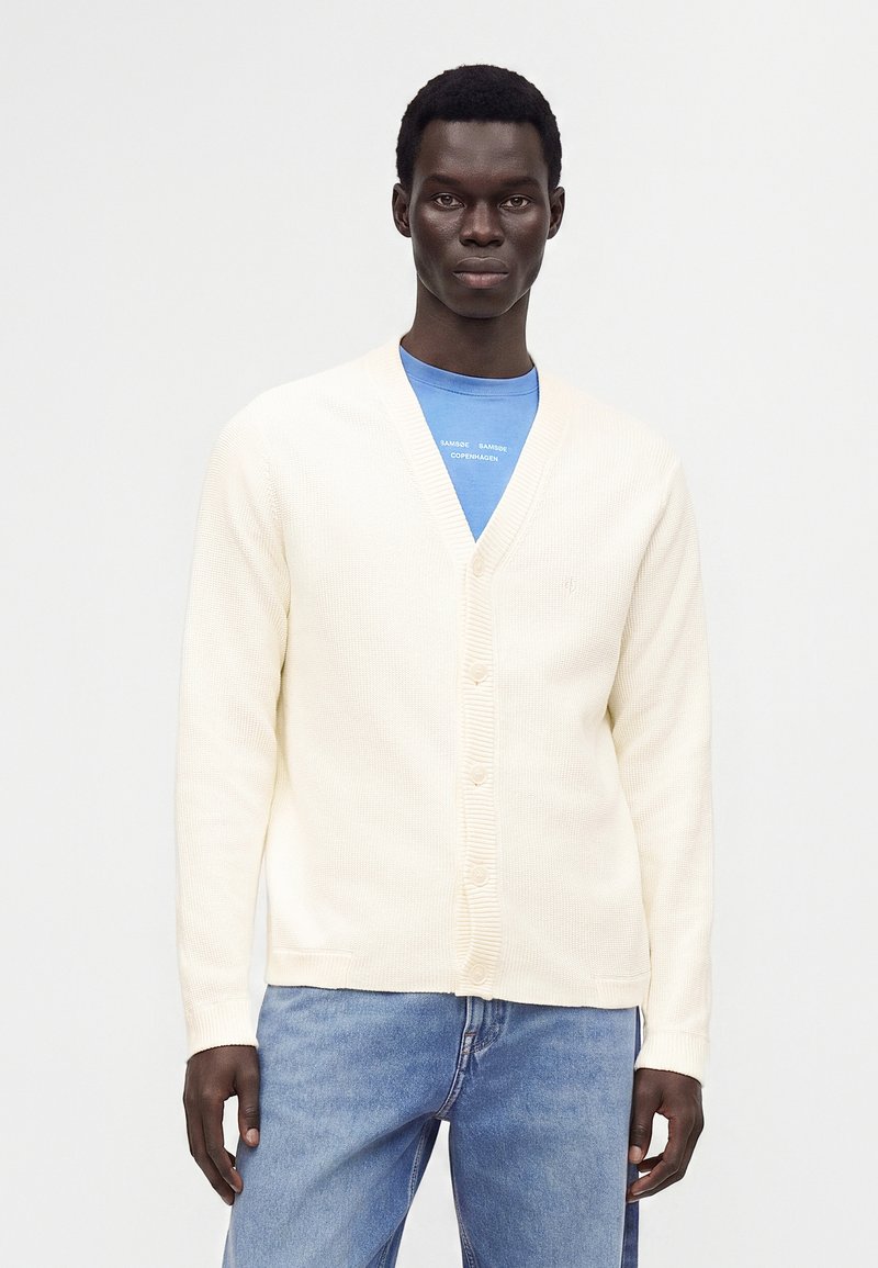 Jeune homme portant un cardigan crème boutonné sur un t-shirt bleu et un jean bleu clair, debout devant un fond blanc uni.