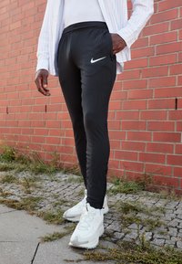 Melnas sporta bikses ar ribotu tekstūru, kurām pa kreisi ir balts Nike logotips, pārī ar baltiem sneakers, stāv uz teksturētas virsmas.