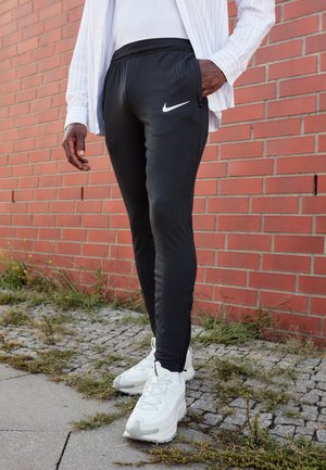 Svarta sportbyxor med ribbad textur, med en vit Nike-logotyp på vänster sida, i kombination med vita sneakers, stående på en texturerad yta.