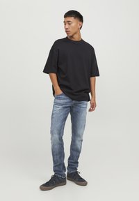 T-shirt nera oversize, jeans in denim blu con una texture sbiadita e sneaker scure. Il modello è in piedi con una mano in tasca, rivolto leggermente di lato.