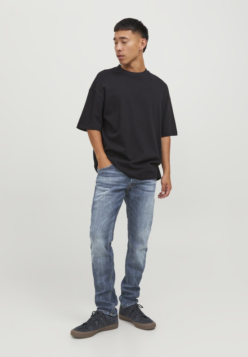 T-shirt nera oversize, jeans in denim blu con una texture sbiadita e sneaker scure. Il modello è in piedi con una mano in tasca, rivolto leggermente di lato.