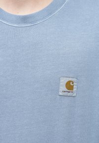Ανοιχτό μπλε βαμβακερό t-shirt με ριμπ λαιμόκοψη και τετράγωνο ετικέτα με κίτρινο λογότυπο και κόκκινο κείμενο "carhartt" στο στήθος.