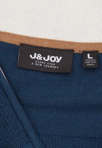 Marineblauw trui met geribbelde textuur, lichtroom en bruine accenten bij de hals, met een label waarop "J&JOY L MADE WITH DIGNITY" staat.