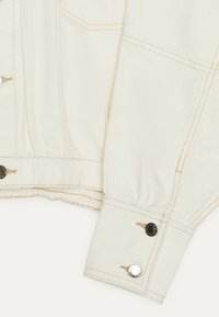 Off-white denim jacket sleeve with fitted cuff, brown stitching, and two metal snap buttons on cuff and waistband.