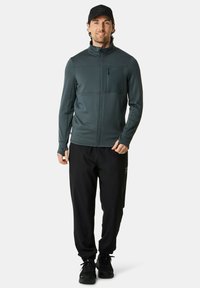 Mørkegrøn zip-up jakke med ribbet tekstur, høj krave og frontlomme, parret med sorte bukser og sorte sneakers.