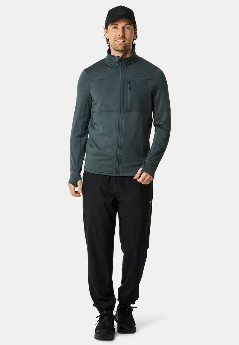 Mørkegrøn zip-up jakke med ribbet tekstur, høj krave og frontlomme, parret med sorte bukser og sorte sneakers.