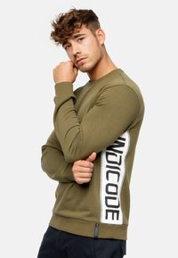 Sweat-shirt vert olive à manches longues, avec une bande latérale blanche portant un texte noir, des poignets côtelés et une petite étiquette de logo à l'ourlet.