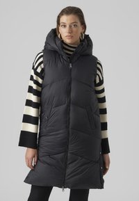 Gilet sans manches noir matelassé avec capuche, doté d'une fermeture éclair à l'avant, de deux poches latérales et d'un motif matelassé en chevrons. Associé à un haut à manches longues rayé.