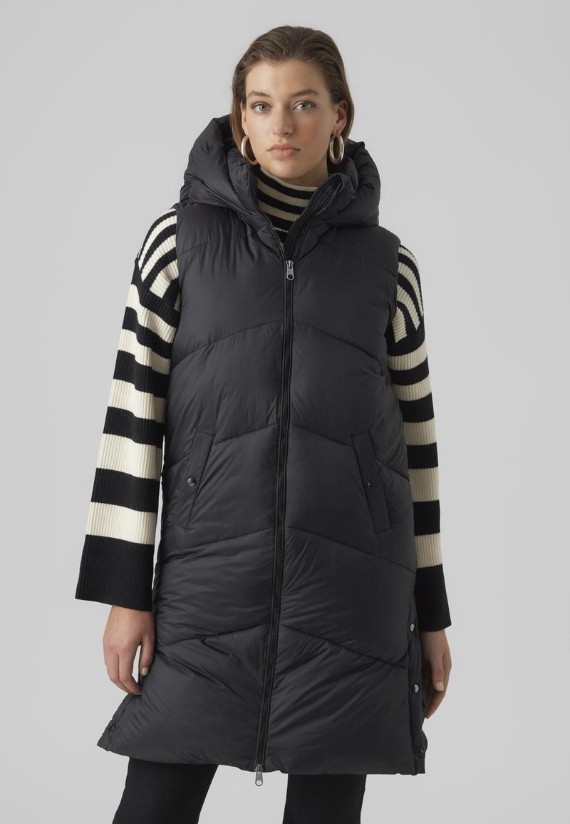 Gilet sans manches noir matelassé avec capuche, doté d'une fermeture éclair à l'avant, de deux poches latérales et d'un motif matelassé en chevrons. Associé à un haut à manches longues rayé.