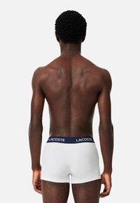 Fehér pamut boxer alsónadrág, navy kék elasztikus derékpánttal, "Lacoste" logóval. A modell hátulról látható, kiemelve a fazont és a kialakítást.