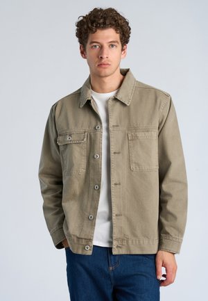 Mand med kort, krøllet hår iført en khakifarvet jakke med knapper over en hvid skjorte og blå jeans, står mod en ensfarvet lys baggrund.
