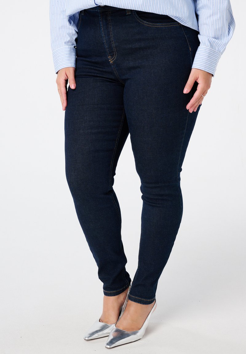 Jean skinny bleu foncé en denim, avec des coutures contrastées, une taille mi-haute et une coupe élégante allant jusqu'à la cheville.