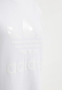 Vit bomullströja med en glänsande vit Adidas-logga, med en stor trefoil-design och text integrerad i tyget.