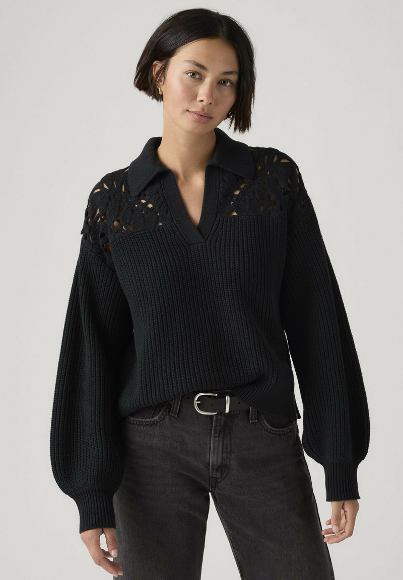 Vrouw met kort donker haar die een zwarte gebreide trui draagt met een kanten inzetstuk, ballonmouwen, een V-hals met kraag, en donkere jeans met een zwarte riem.