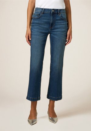 Femme portant un jean court taille mi-haute bleu foncé avec des talons pointus argentés et une chemise blanche rentrée, debout devant un fond uni.