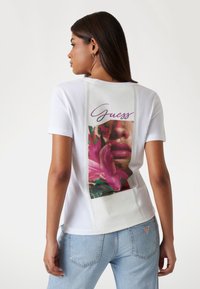 Balta marškinėliai su gėlių grafika ir "Guess" tekstu ant nugaros. Pagaminti iš medvilnės, prigludęs dizainas ir kontrastingos siūlės detalė.