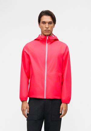 Άνδρας που φοράει φωτεινό κόκκινο μπουφάν με κουκούλα, εμπρόσθιο φερμουάρ και πλαϊνές τσέπες, στέκεται μπροστά σε ένα απλό γκρι φόντο.