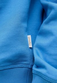 Artimas mėlynos sportinės palaidinės šono siūlės vaizdas, rodyti mažą baltą audinio etiketę su kursyvine tekstu.