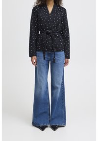 Top noir à enrouler avec motif de cœurs blancs, noué à la taille, associé à un jean bleu flare taille haute et des escarpins noirs à bout pointu.