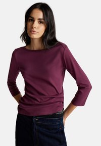 T-shirt bordeaux à manches trois-quarts en tissu doux avec un col rond et une texture lisse, associé à un jean foncé avec une taille visible.