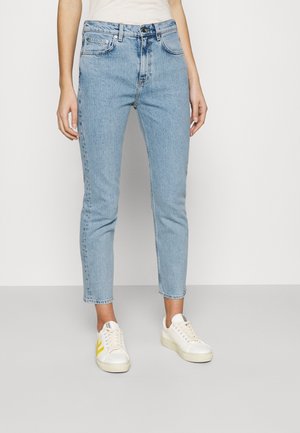 Person, die hellblaue Slim-Fit-Jeans und weiße Sneakers mit gelben Akzenten trägt, steht vor einem einfarbigen hellen Hintergrund.