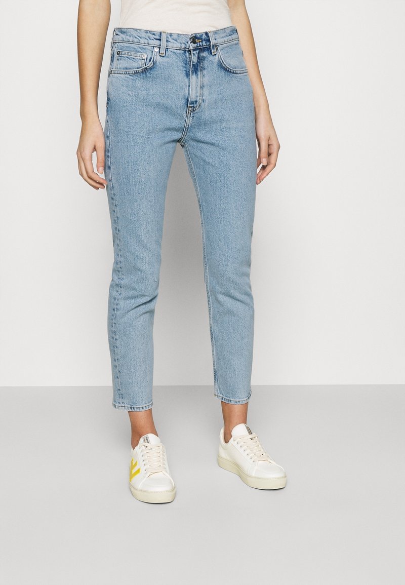 Personne portant un jean slim bleu clair et des baskets blanches avec des touches jaunes, debout devant un fond clair uni.