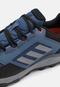 Chaussure de sport bleue et noire avec une tige en maille, des rayures grises et des accents orange. Semelle en caoutchouc texturé pour une meilleure adhérence.