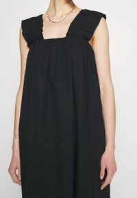 Černé bezrukávové šaty se čtvercovým výstřihem, nařaseným tělem a volánkovými ramínky, vyrobené z měkké, lehké látky.