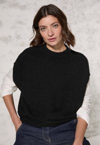 Sort strikket sweater med en tekstureret overflade, oversized pasform og rund hals. Bæres over en hvid skjorte med krave, parret med blå jeans.