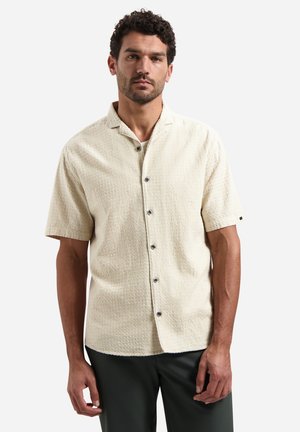 Mann mit lockigem Haar, der ein cremefarbenes, strukturiertes Kurzarmhemd mit Knöpfen und dunkle Hosen trägt, steht vor einem schlichten hellen Hintergrund.