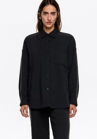 Camisa negra, ligera y texturizada, con cierre de botones en la parte delantera, mangas largas, cuello y bolsillo en el pecho. Combinada con pantalones a juego.